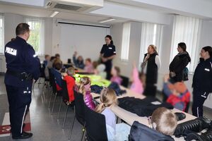Policjanci oprowadzają dzieci po dziedzińcu komendy, pokazują radiowozy oraz prowadzą spotkanie na sali odpraw, podczas którego dzieci korzystają z zabawek edukacyjnych (samochodów i drogi z przejściem dla pieszych)