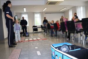 Policjanci oprowadzają dzieci po dziedzińcu komendy, pokazują radiowozy oraz prowadzą spotkanie na sali odpraw, podczas którego dzieci korzystają z zabawek edukacyjnych (samochodów i drogi z przejściem dla pieszych)