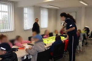 Policjanci oprowadzają dzieci po dziedzińcu komendy, pokazują radiowozy oraz prowadzą spotkanie na sali odpraw, podczas którego dzieci korzystają z zabawek edukacyjnych (samochodów i drogi z przejściem dla pieszych)