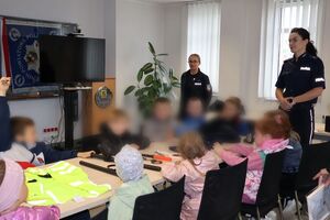 Policjanci oprowadzają dzieci po dziedzińcu komendy, pokazują radiowozy oraz prowadzą spotkanie na sali odpraw, podczas którego dzieci korzystają z zabawek edukacyjnych (samochodów i drogi z przejściem dla pieszych)