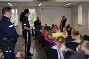 Policjanci oprowadzają dzieci po dziedzińcu komendy, pokazują radiowozy oraz prowadzą spotkanie na sali odpraw, podczas którego dzieci korzystają z zabawek edukacyjnych (samochodów i drogi z przejściem dla pieszych)