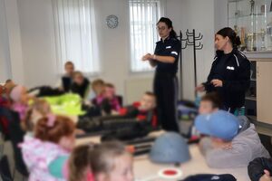 Policjanci oprowadzają dzieci po dziedzińcu komendy, pokazują radiowozy oraz prowadzą spotkanie na sali odpraw, podczas którego dzieci korzystają z zabawek edukacyjnych (samochodów i drogi z przejściem dla pieszych)