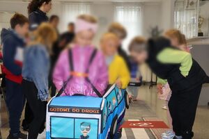 Policjanci oprowadzają dzieci po dziedzińcu komendy, pokazują radiowozy oraz prowadzą spotkanie na sali odpraw, podczas którego dzieci korzystają z zabawek edukacyjnych (samochodów i drogi z przejściem dla pieszych)