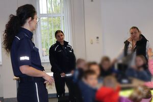 Policjanci oprowadzają dzieci po dziedzińcu komendy, pokazują radiowozy oraz prowadzą spotkanie na sali odpraw, podczas którego dzieci korzystają z zabawek edukacyjnych (samochodów i drogi z przejściem dla pieszych)