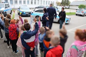 Policjanci oprowadzają dzieci po dziedzińcu komendy, pokazują radiowozy oraz prowadzą spotkanie na sali odpraw, podczas którego dzieci korzystają z zabawek edukacyjnych (samochodów i drogi z przejściem dla pieszych)