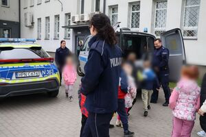 Policjanci oprowadzają dzieci po dziedzińcu komendy, pokazują radiowozy oraz prowadzą spotkanie na sali odpraw, podczas którego dzieci korzystają z zabawek edukacyjnych (samochodów i drogi z przejściem dla pieszych)