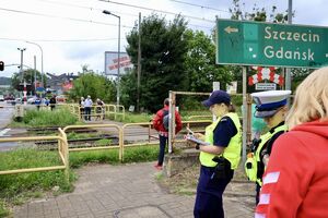 Policjanci ze strażnikami ochrony kolei rozmawiają z kierowcami, którzy zatrzymali się przed przejazdem kolejowym na zamkniętych rogatkach.