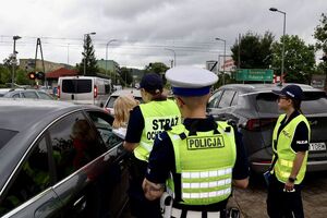 Policjanci ze strażnikami ochrony kolei rozmawiają z kierowcami, którzy zatrzymali się przed przejazdem kolejowym na zamkniętych rogatkach.