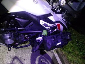 Policjanci pracują na miejscu wypadków, na zdjęciu widoczny jest niebieski samochód z rozbitym przodem, motocykl, rowerzysta przejeżdżający przed maską samochodu oraz radiowóz postawiony za białym samochodem oraz w innym miejscu radiowóz stojący przy chodniku.