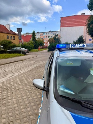 Policjanci pracują na miejscu wypadków, na zdjęciu widoczny jest niebieski samochód z rozbitym przodem, motocykl, rowerzysta przejeżdżający przed maską samochodu oraz radiowóz postawiony za białym samochodem oraz w innym miejscu radiowóz stojący przy chodniku.